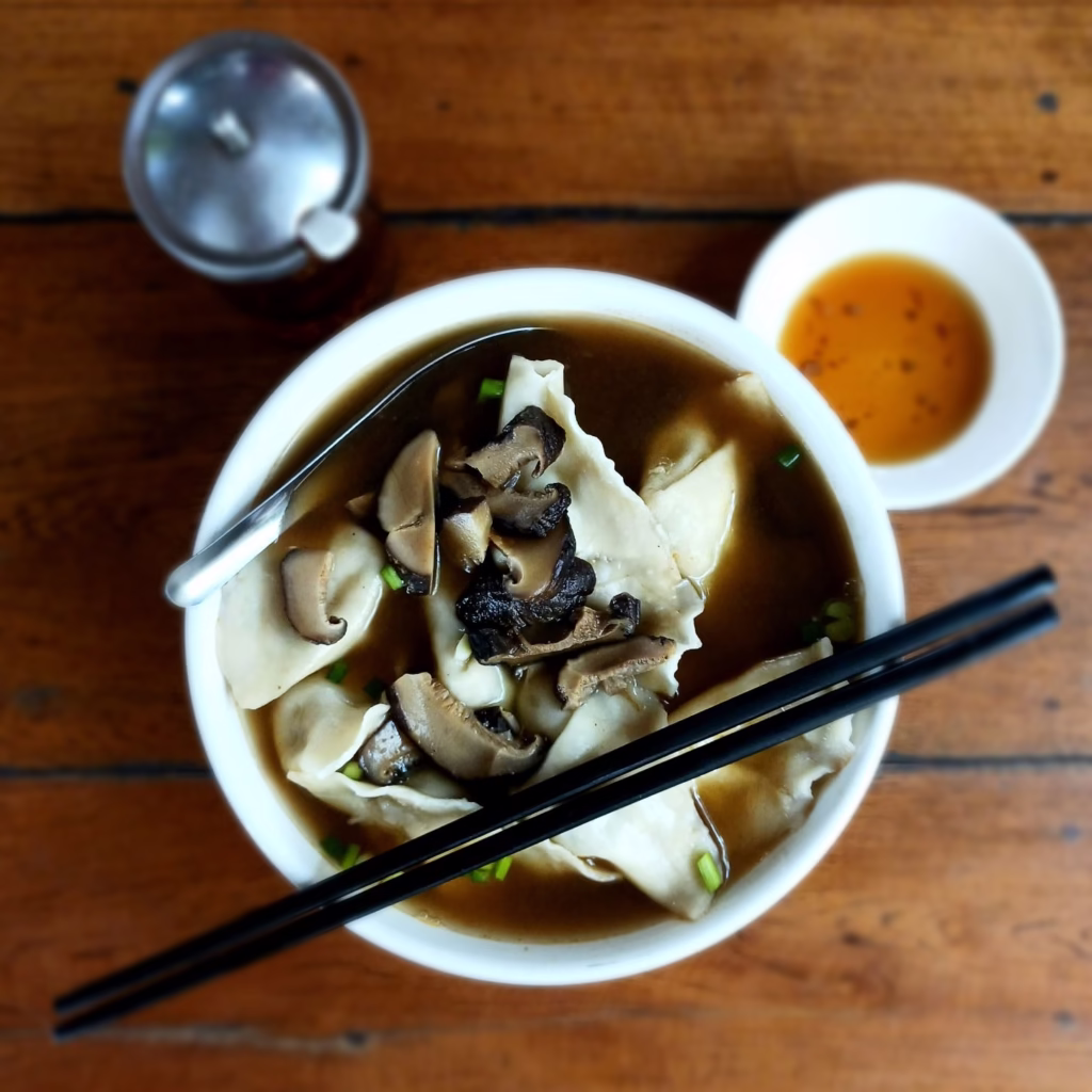 Brown broth with 10 large vegan dumplings and mushrooms. Chopsticks place over the top of the bowl. Chili sauce in a small saucer in the top right